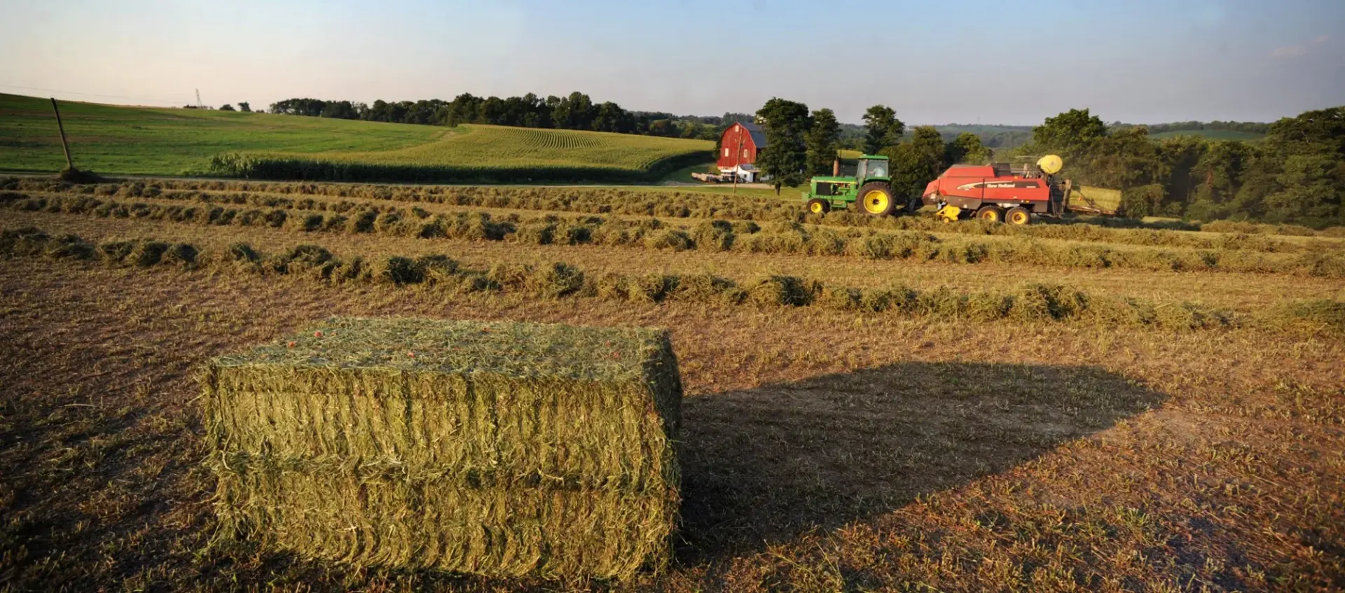 A tractor and baler working a hay field below a red barn on the hill